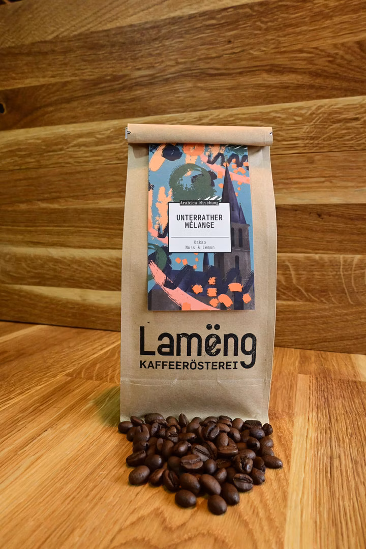 Lamëng-Kraftpapier-Tüte mit Unterrather-Melange-Etikett auf Holzboden, davor verschüttete frische Kaffeebohnen