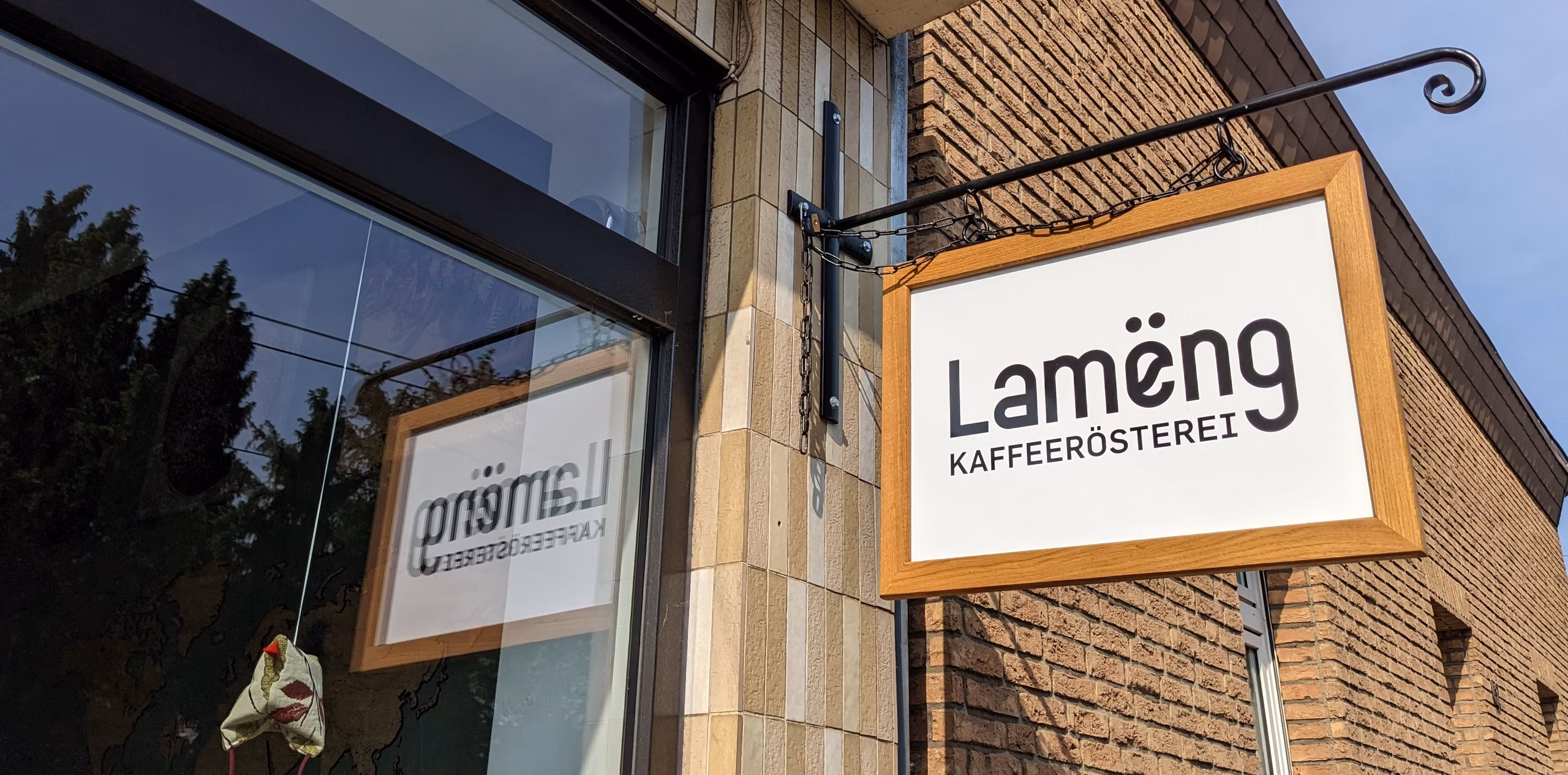 Aussenfassade der Rösterei Lamëng in Düsseldorf-Unterrath, das geschwungene Café-Schild „Lamëng KAFFEERÖSTEREI" hängt seitlich an einem schwarzen Schmiedeeisen-Ausleger über dem Eingang